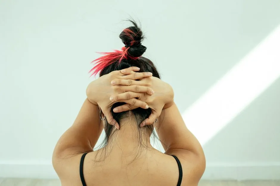Back view of anonymous female stretching neck pressing clasped hands on head against white wall
