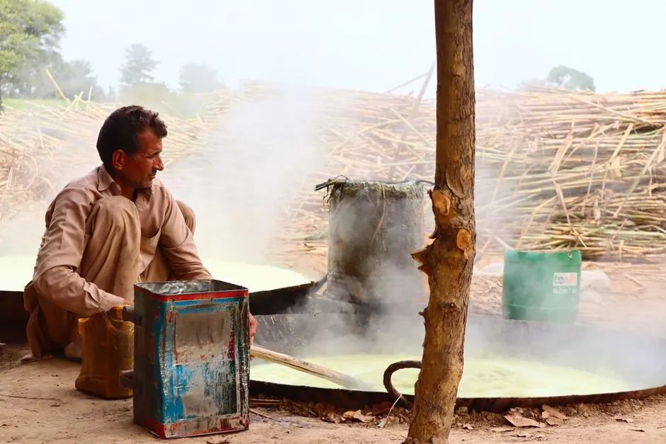A man boiling sugarcane juice outdoors with steam rising in a rural setting.