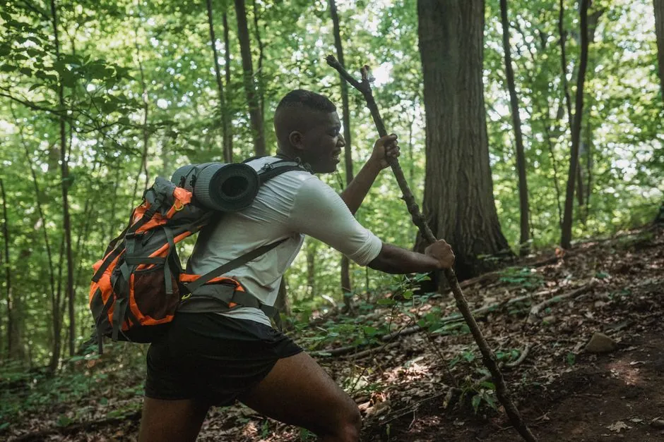 Side view of black young man with backpack climbing with tree branch among green woods on sunny summer day