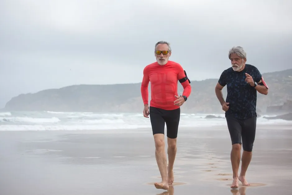 Two senior men jogging barefoot by the ocean for fitness on a cloudy beach in Portugal.
