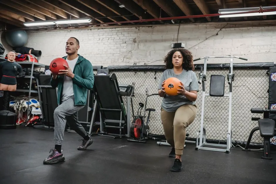 Full body of focused overweight African American female with fit ethnic male trainer doing forward lunges with medicine balls during functional workout in gym