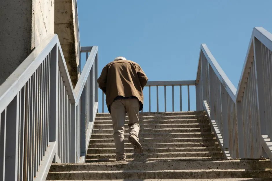 A senior man climbs a set of concrete steps beneath a clear blue sky, captured from behind.