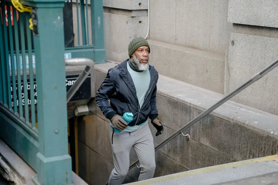 Concentrated senior African American male in windbreaker jacket and sweatpants going upstairs with fitness bottle in hand