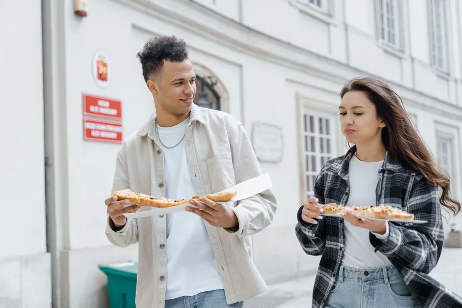 Two young adults enjoying pizza while walking outdoors in the city, creating a joyful street food moment.