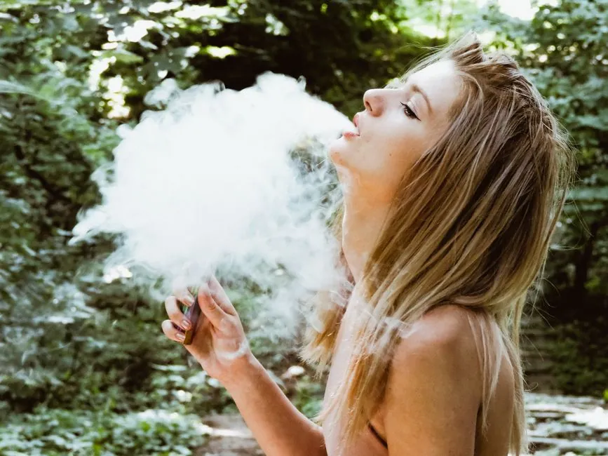 Portrait of a young woman vaping in a lush outdoor setting on a sunny day.