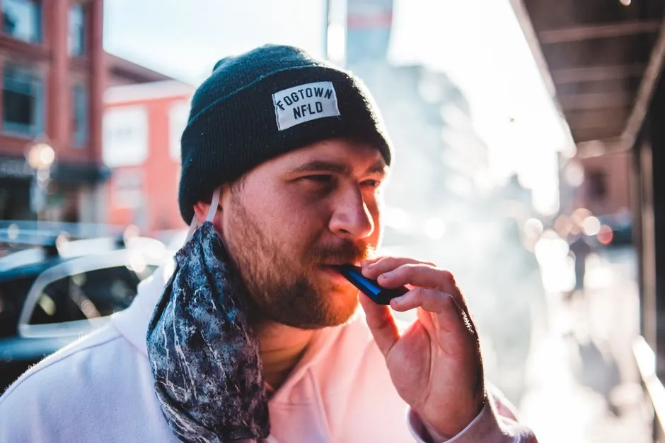 Casual young man vaping outdoors in a city street, enjoying a sunny day.