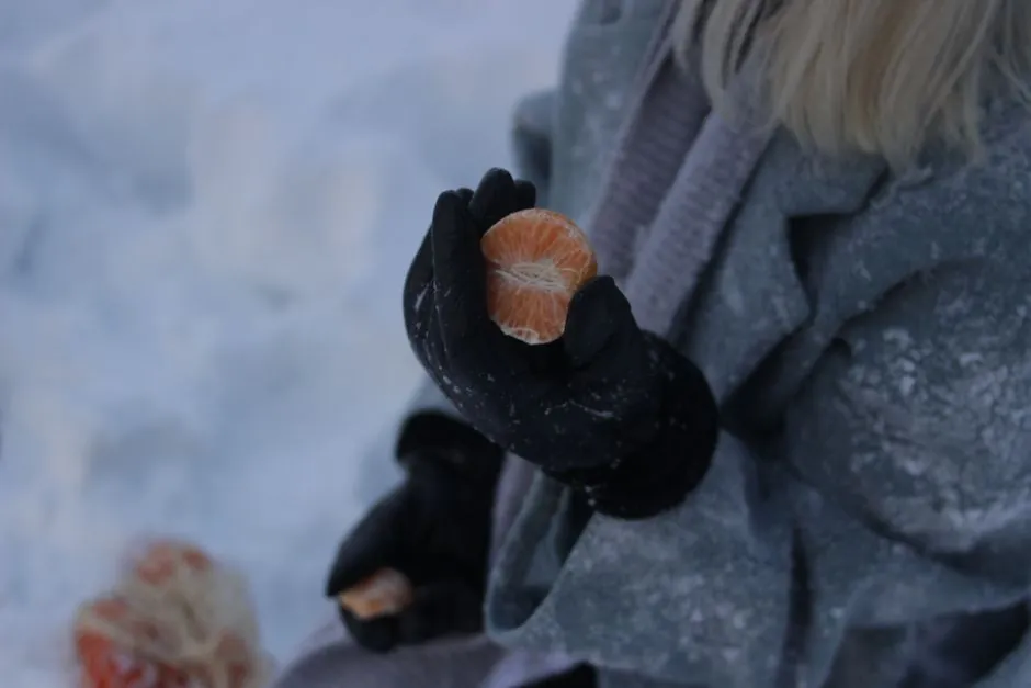 Close-up of a person holding a tangerine slice in winter gloves outdoors.