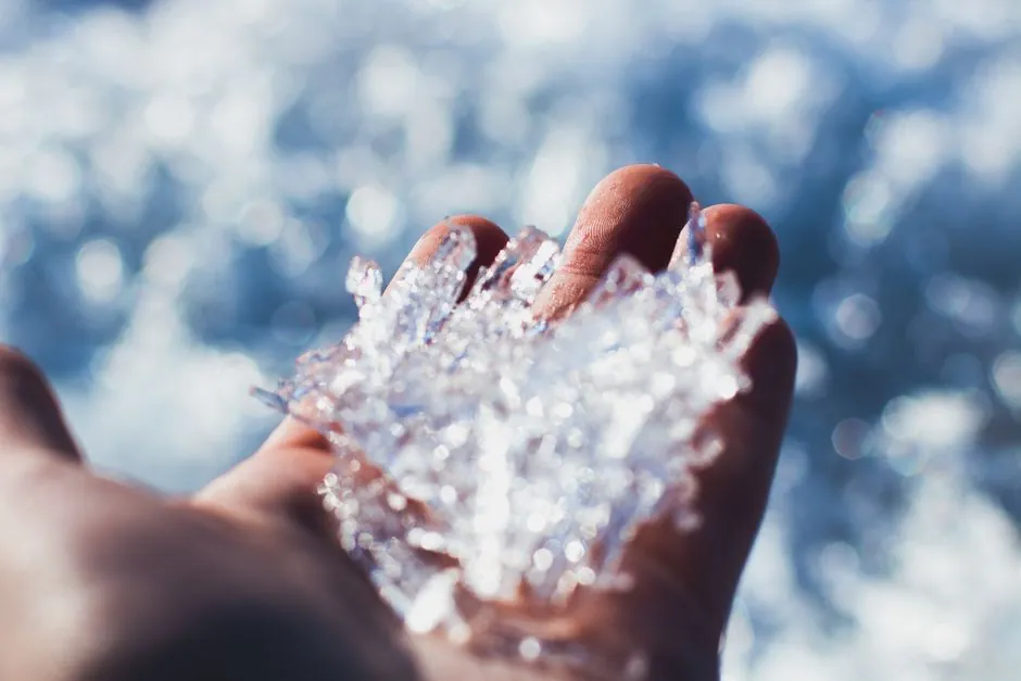 From above of crop anonymous person demonstrating piece of textured ice in hand against blurred white snowy terrain on sunny day