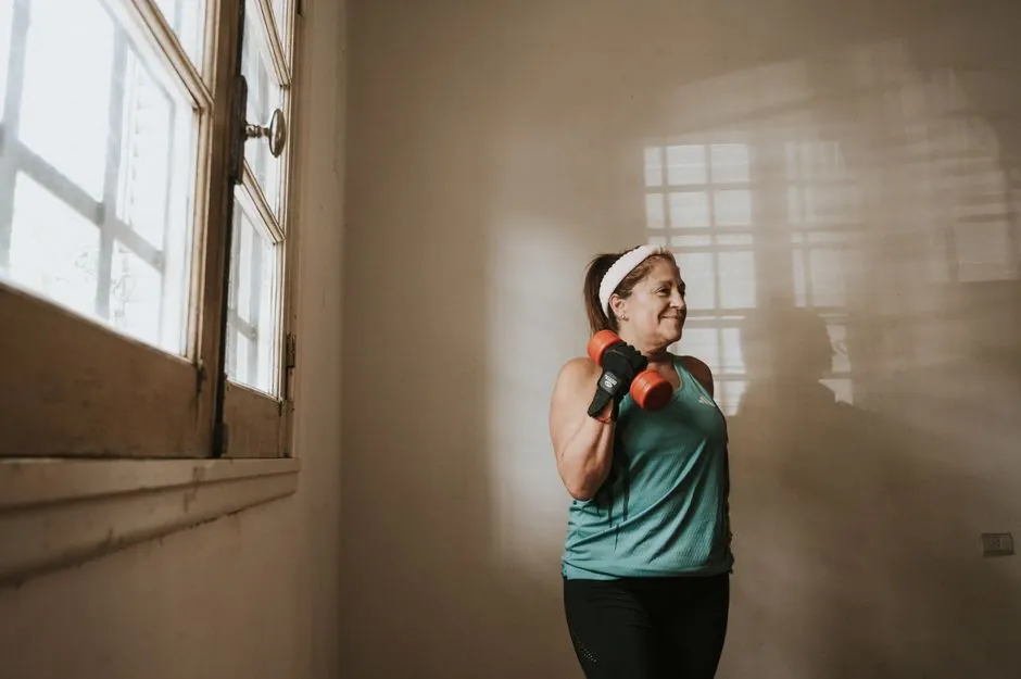 Senior woman in a brightly lit room exercises with dumbbells, promoting fitness at home.