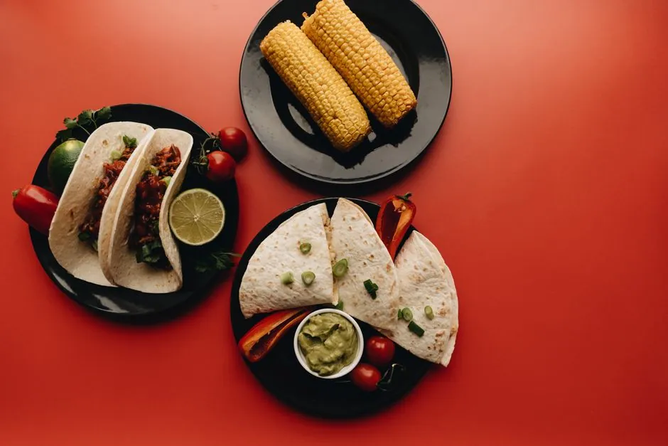Top view of Mexican dishes including tacos, corn, and quesadillas on vibrant red background.