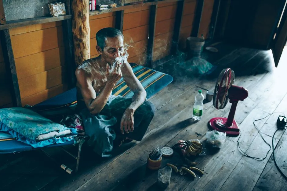 An elderly man sits on a cot, smoking, in a traditional wooden interior space with natural light.