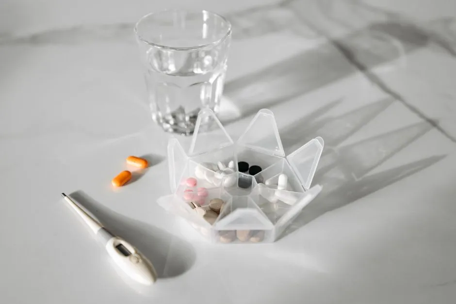 A pill organizer with medication, a glass of water, and a thermometer on a white surface.