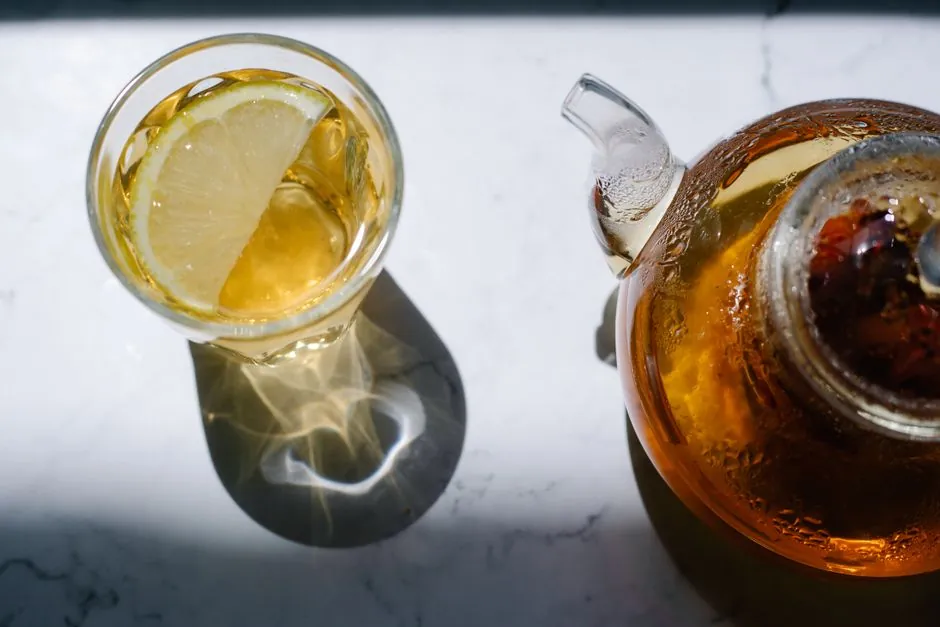 Top view of lemon tea in a glass alongside a teapot on a marble surface, highlighted in natural light.