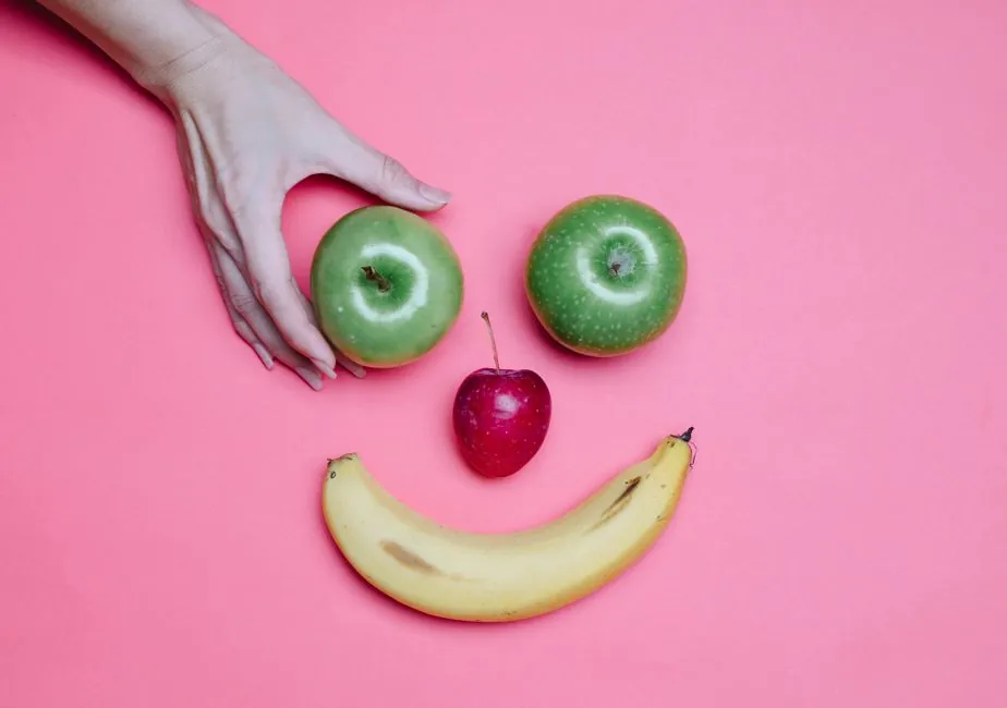Top view of crop anonymous female making smiling face out of apples and banana on pink background